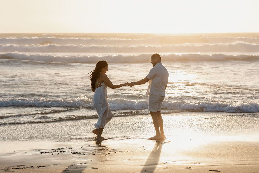 A couple holds hands and dances playfully together on a sunlit beach during golden hour.