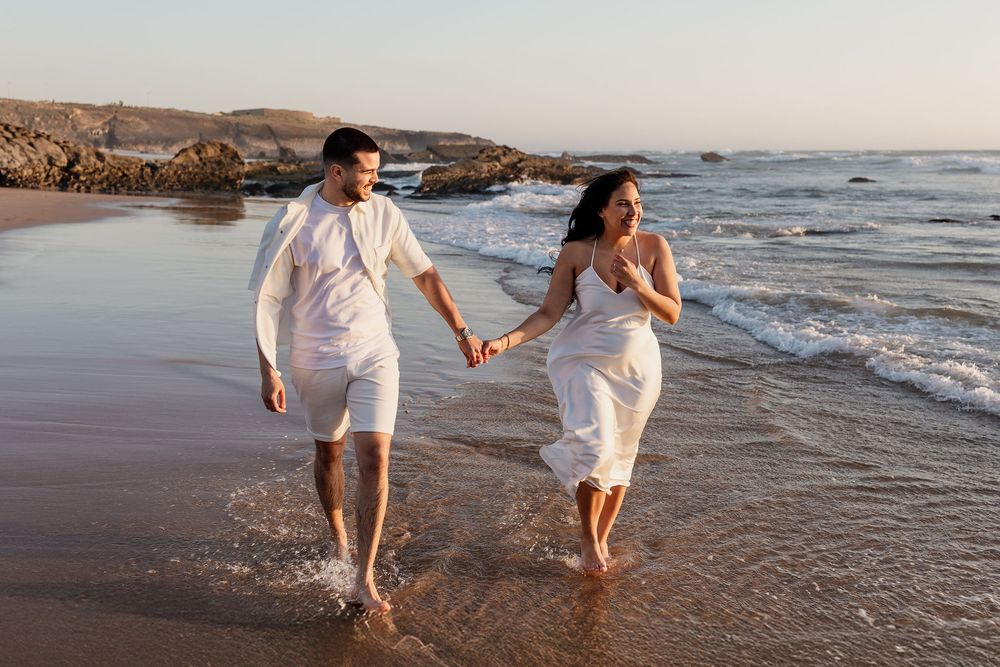 A couple in white outfits runs playfully along the beach at sunset, splashing through the ocean waves.
