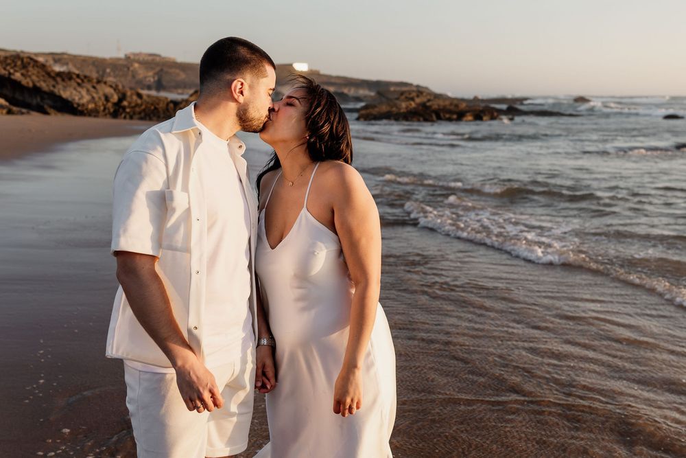 A romantic couple in white outfits sharing an intimate moment on a beach at sunset.