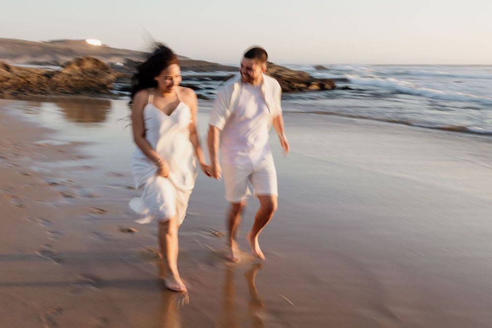 A couple in white clothing runs joyfully along a beach at sunset, creating a romantic seaside moment.