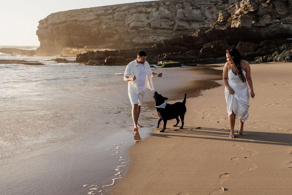 Two people in white outfits walking a black dog on a scenic beach with rocky cliffs during golden hour sunset.