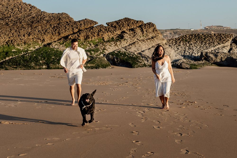 A couple in white outfits runs with their black dog on a sandy beach with cliffs in the background.