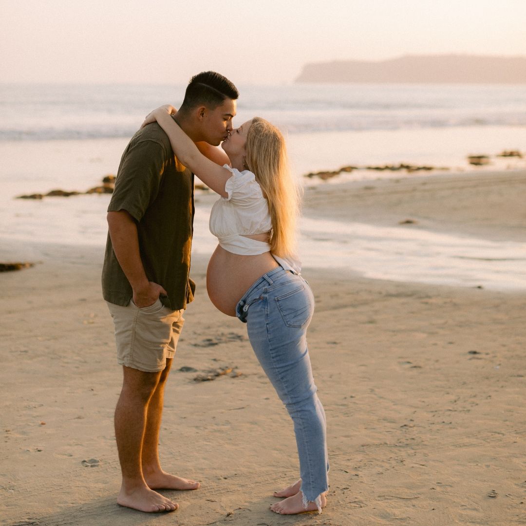 A couple shares a romantic moment on a misty beach during sunset, standing barefoot in the sand. Maternity photoshoot San Diego