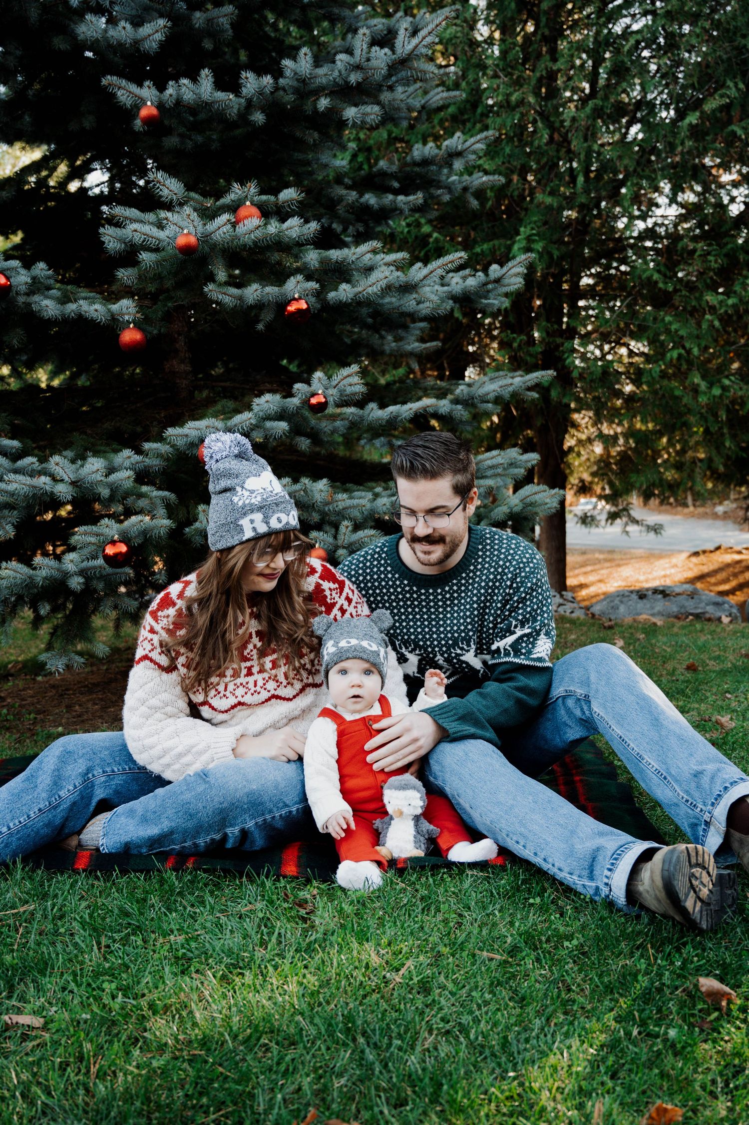 Family wearing cozy winter sweaters sits together on grass near decorated Christmas tree outdoors.