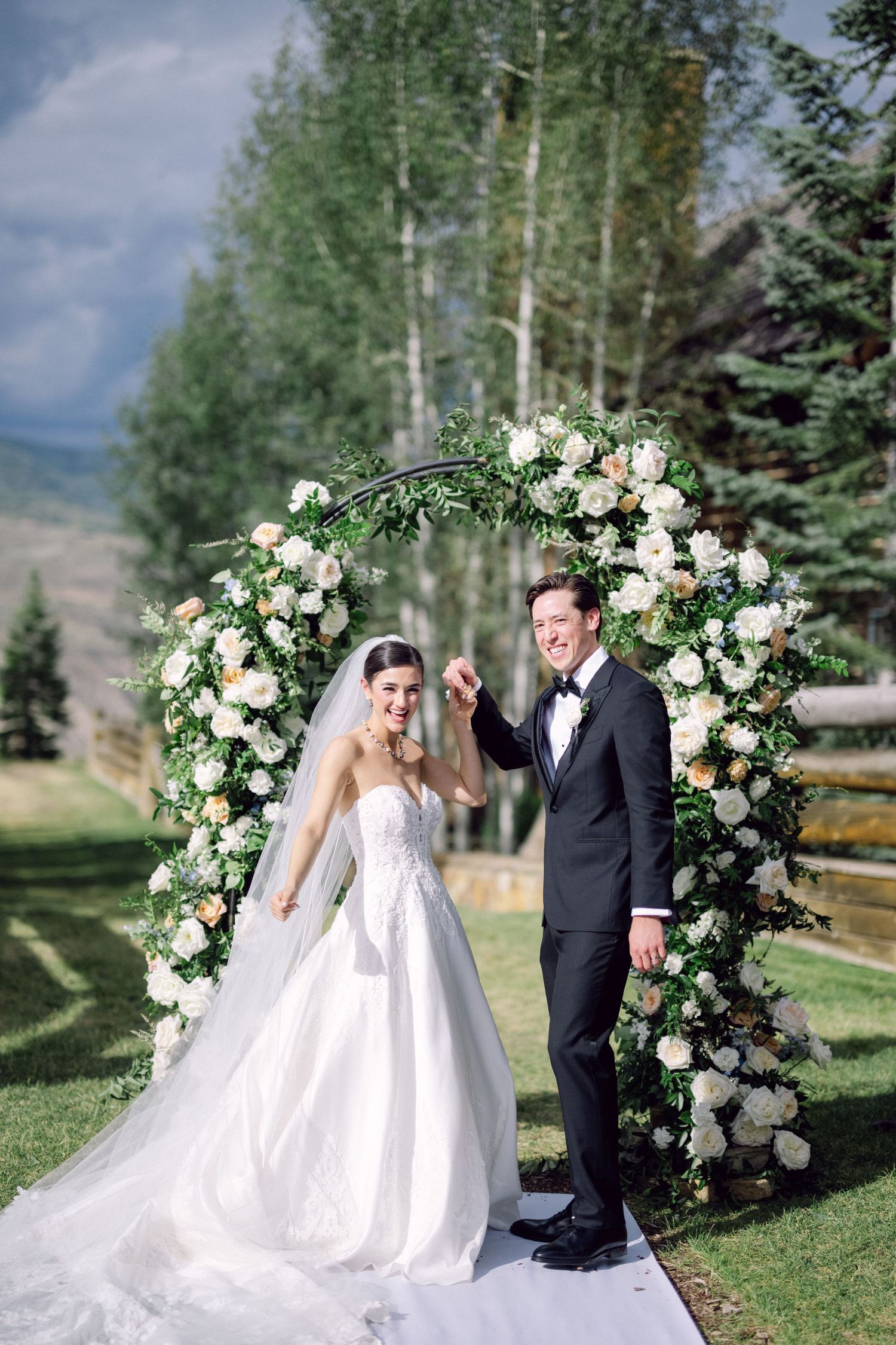 Newlyweds standing under an elegant floral arch with white roses against a mountain backdrop.