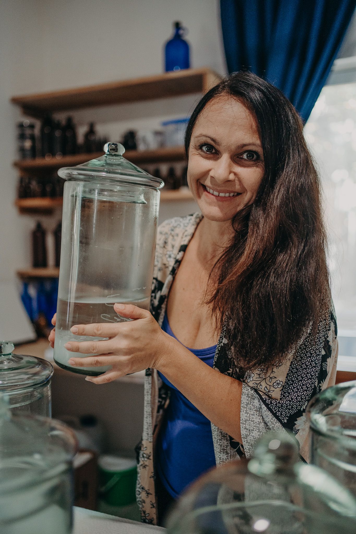 A woman holds a large glass jar in a kitchen while wearing a patterned dress with navy curtains and shelves in background.