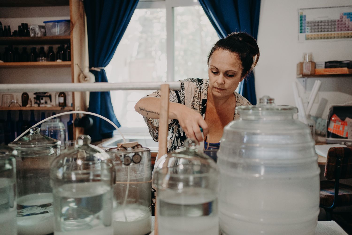 Person preparing beverages with multiple glass jars and containers on a counter near blue curtains.
