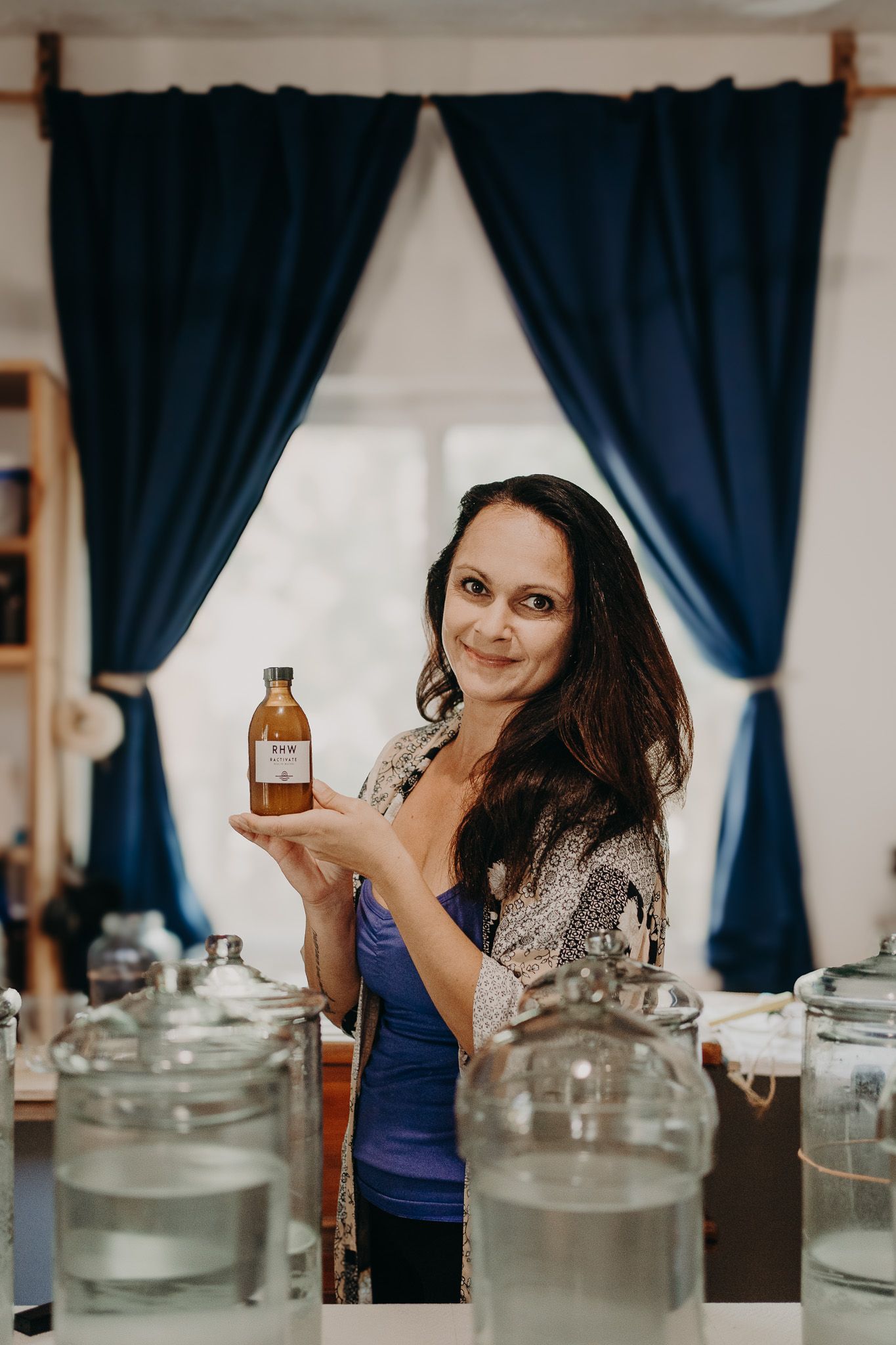 Person holding a brown bottle among various glass containers and blue curtains in background.