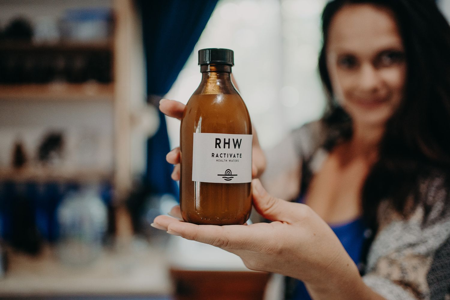 Hand holding a brown bottle labeled RHW against a blurred indoor background.