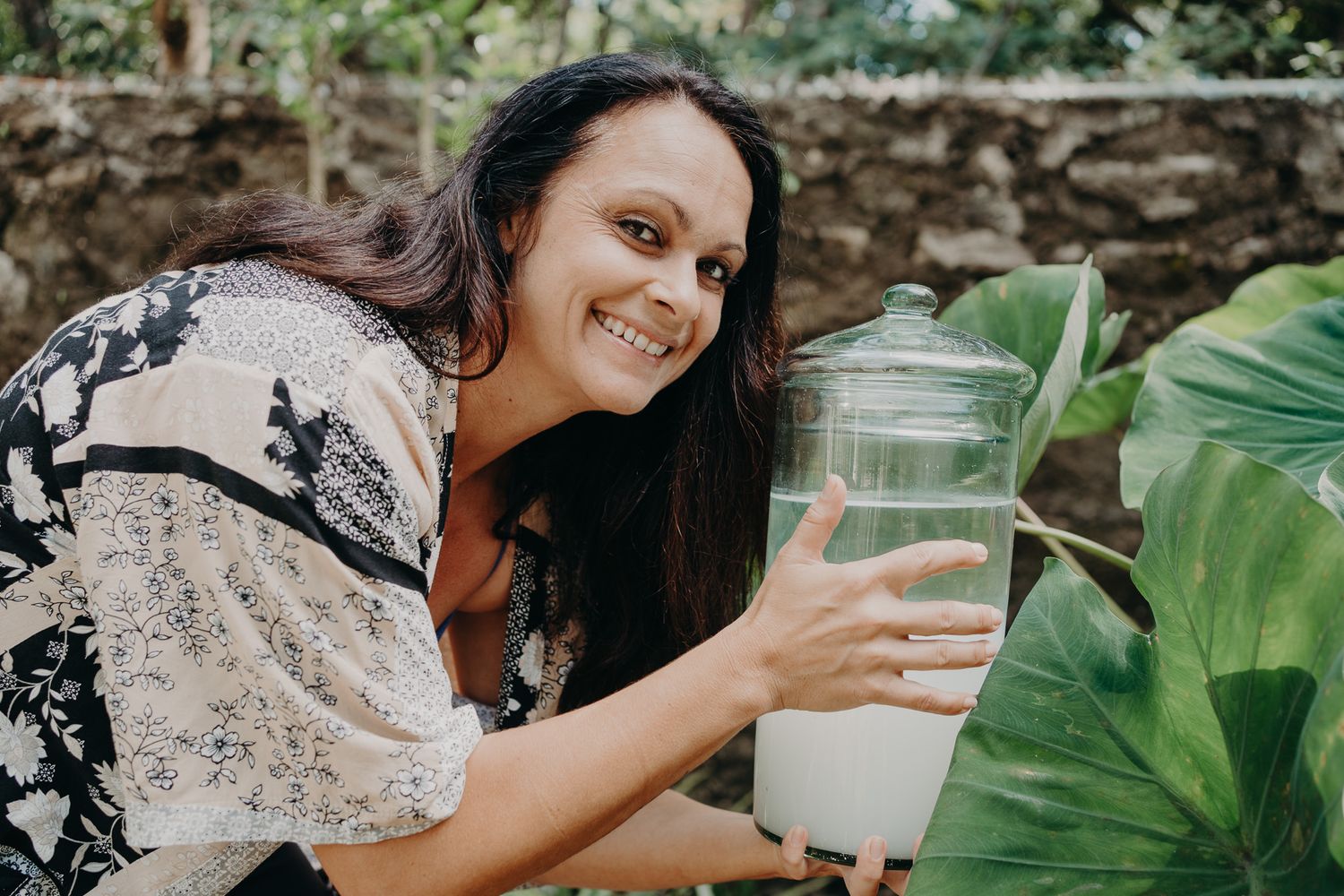 Person with glass container among tropical plants and greenery outdoors.