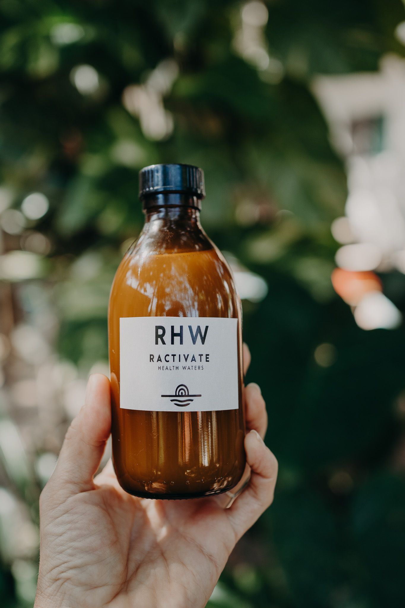 Hand holding brown RHW branded bottle against bokeh nature background.