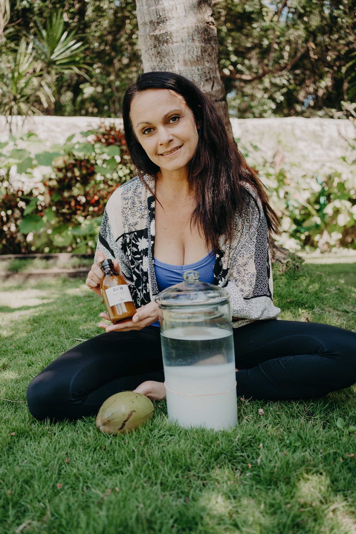 Person sitting on grass holding a bottle and jar in an outdoor garden setting.