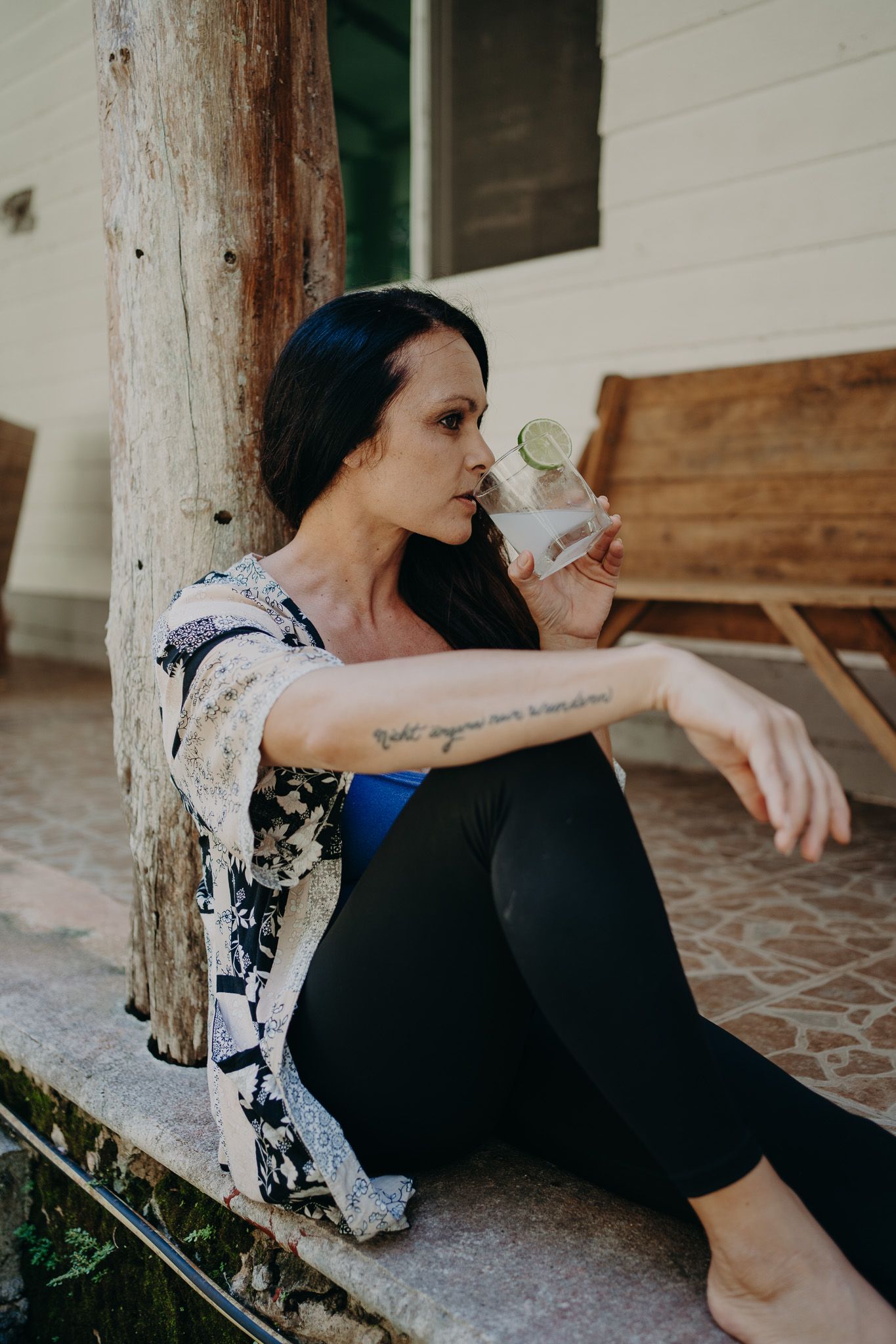 Person in floral kimono and black pants sitting by wooden post drinking from glass photoshoot in Puerto Morelos.