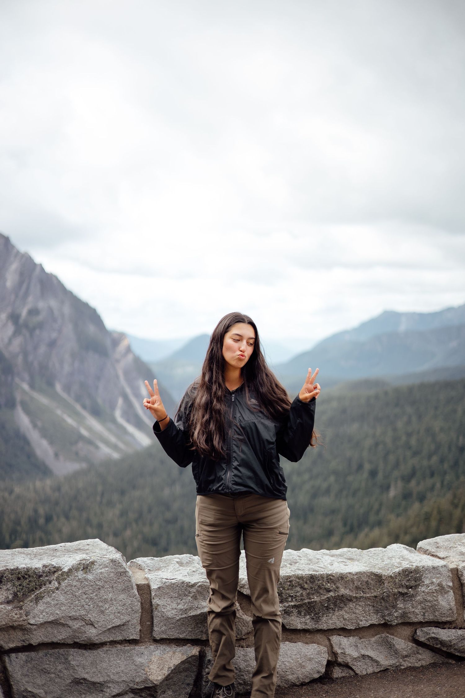 A person stands on a stone wall making peace signs with scenic mountain views in Banff National Park, Canada.