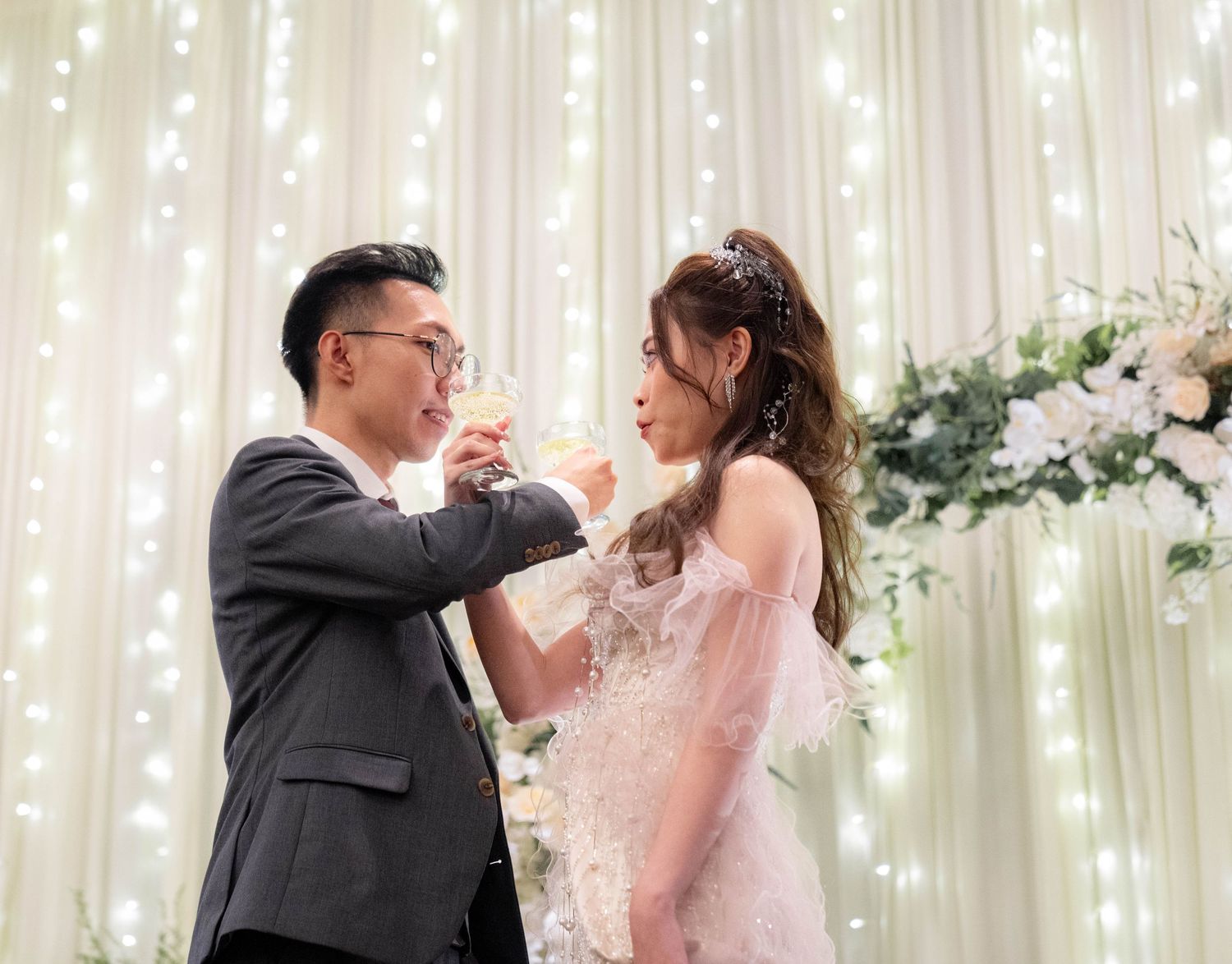 A couple in wedding attire share a playful moment together on a lawn with white buildings in the background.