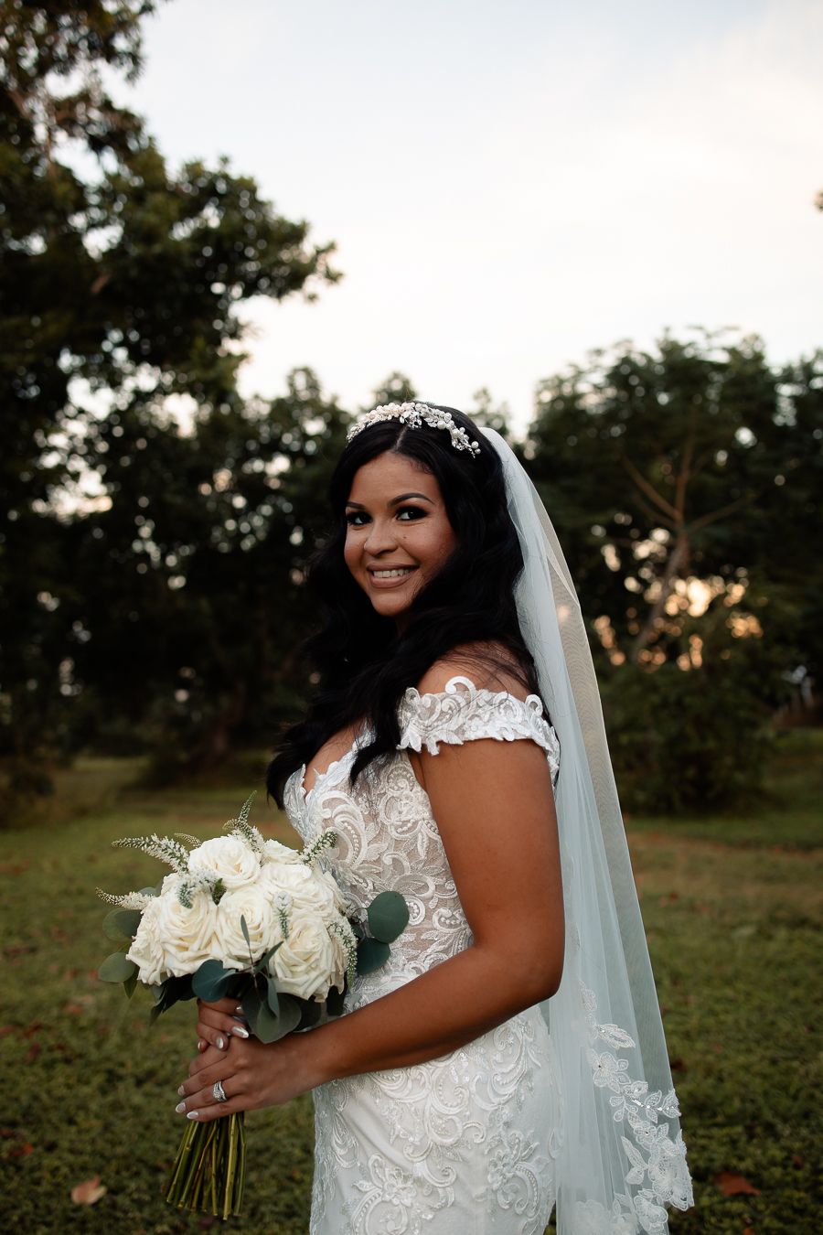 Beautiful bride in lace gown holds white rose bouquet during outdoor wedding photoshoot.