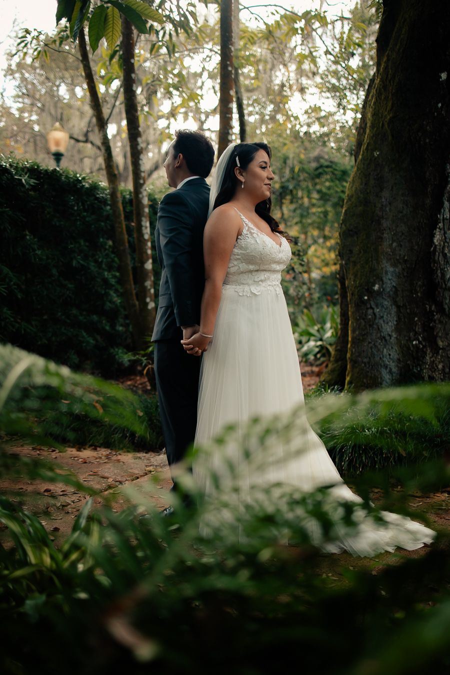 Bride and groom holding hands, standing back-to-back outdoors, surrounded by greenery.