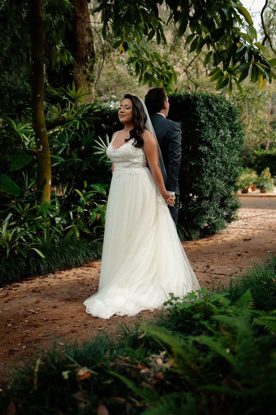 Bride and groom standing back-to-back, holding hands before their first look in a lush garden.