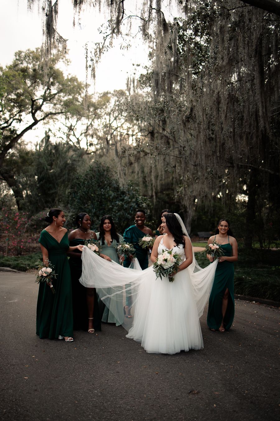 Bride surrounded by bridesmaids in green dresses, holding her train and bouquet outdoors.