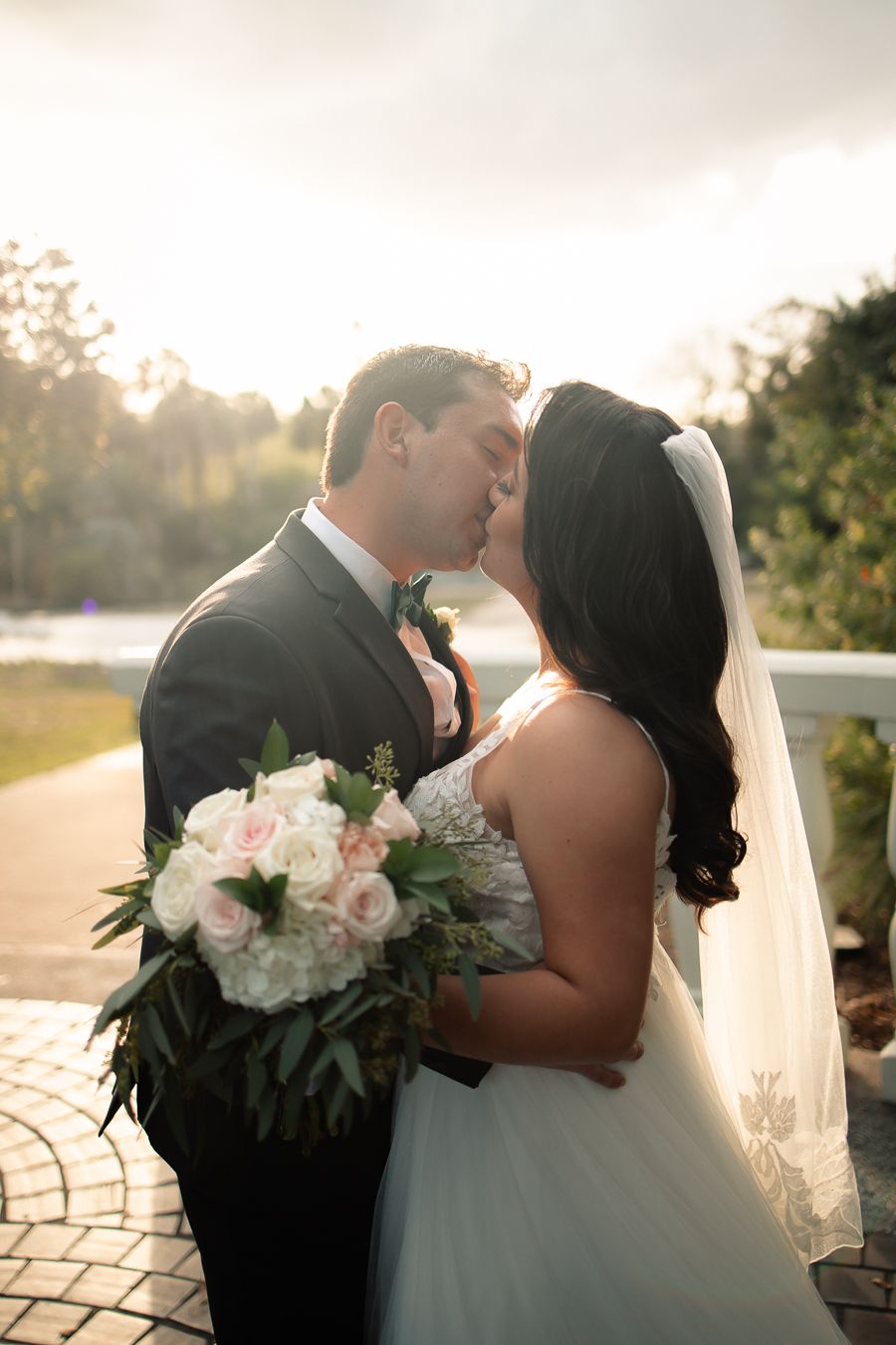 Romantic sunset portrait of newlyweds sharing a kiss while holding blush pink bouquet.