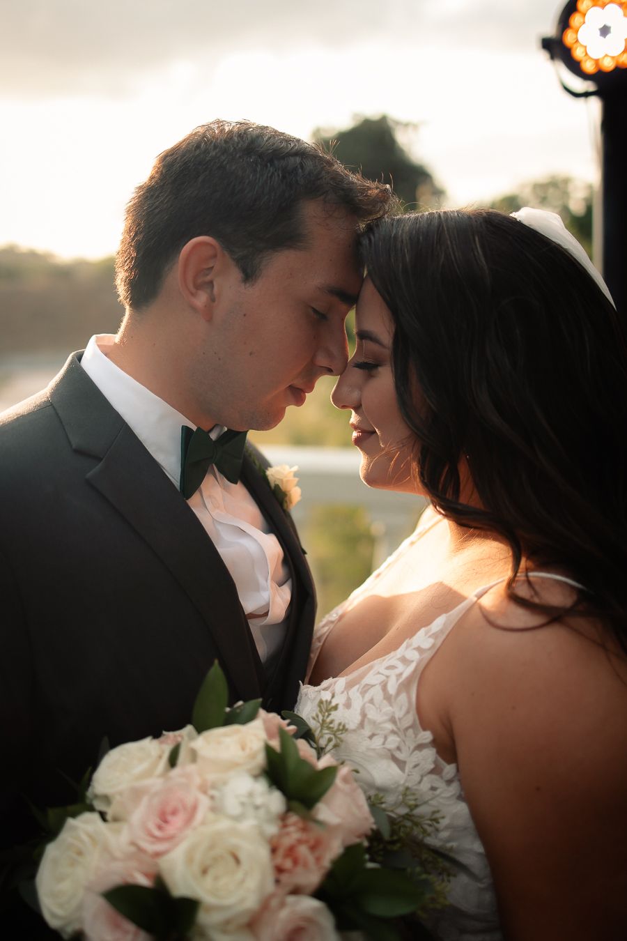 Bride and groom forehead-to-forehead, holding a bouquet with soft sunset lighting.