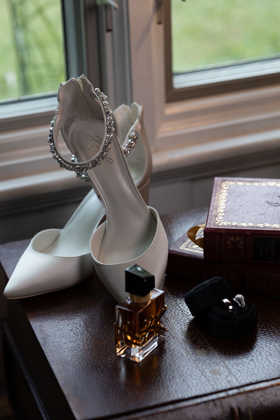 White bridal shoes, perfume bottle, and ring box arranged on wooden windowsill.