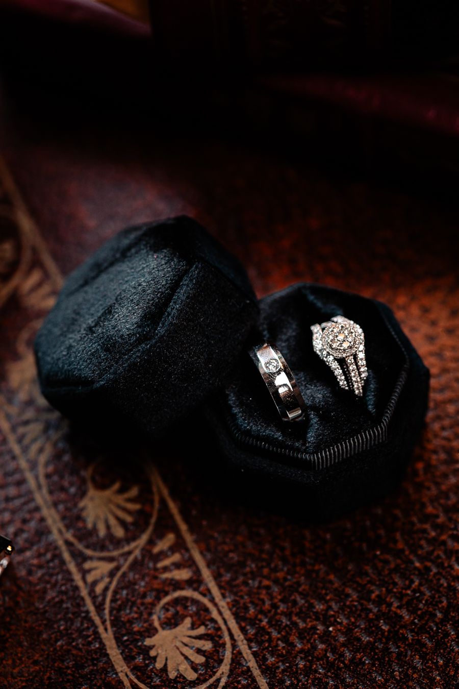 Diamond wedding rings nestled in black velvet ring box on patterned surface.