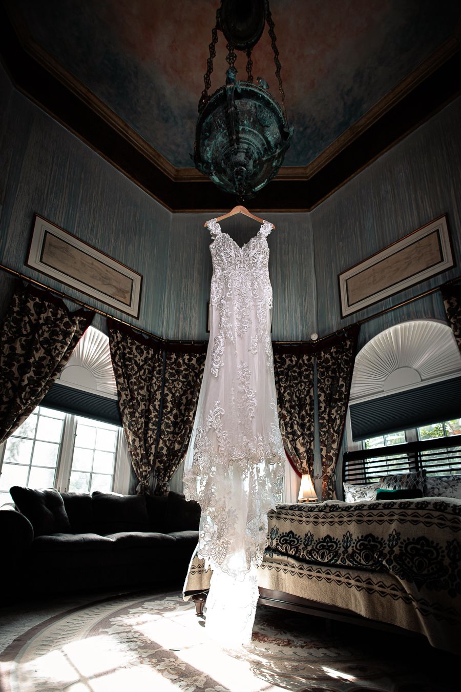 Lace wedding dress hanging dramatically from chandelier in elegant room with natural lighting.