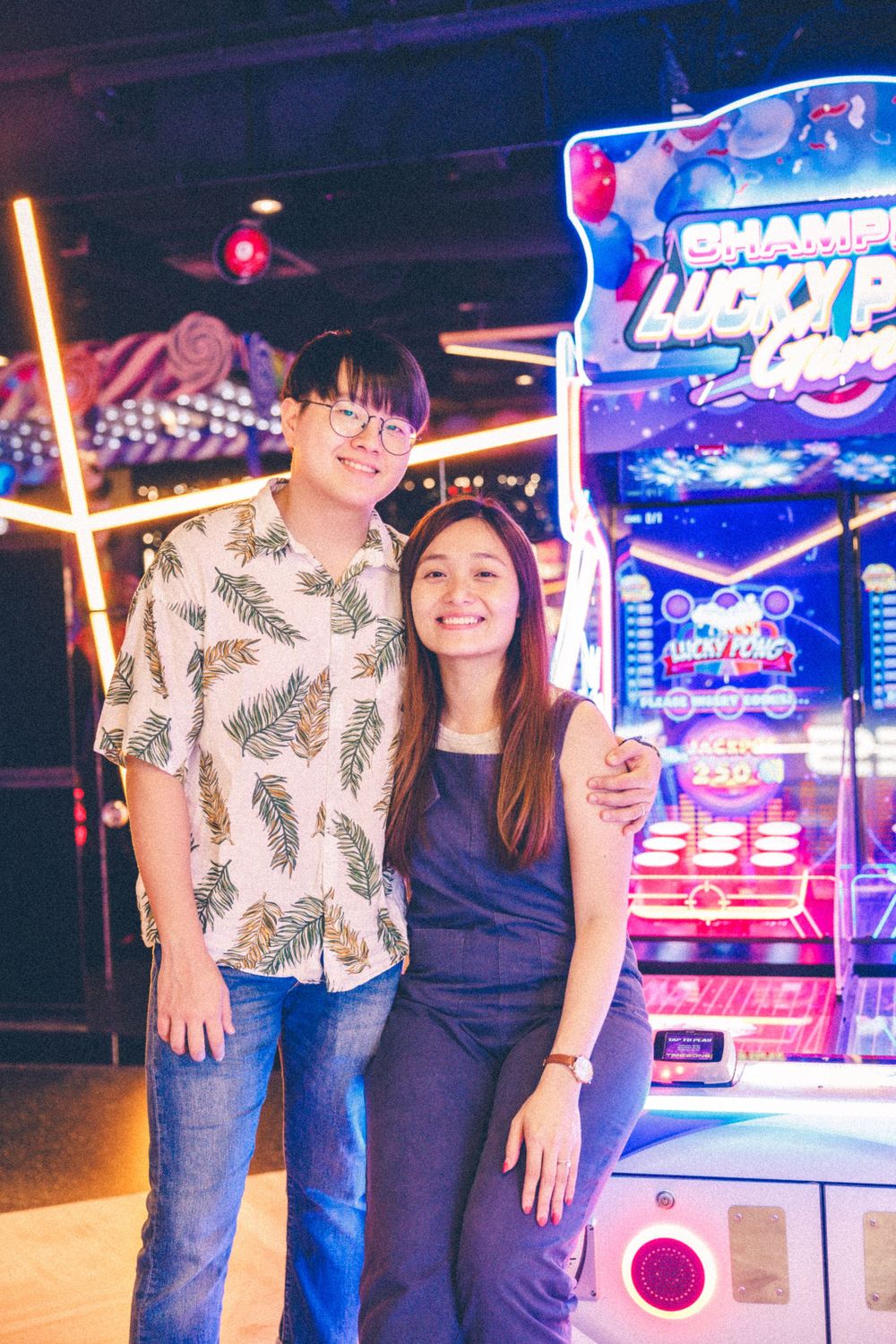 A couple poses together beside an illuminated arcade game machine with neon signs.