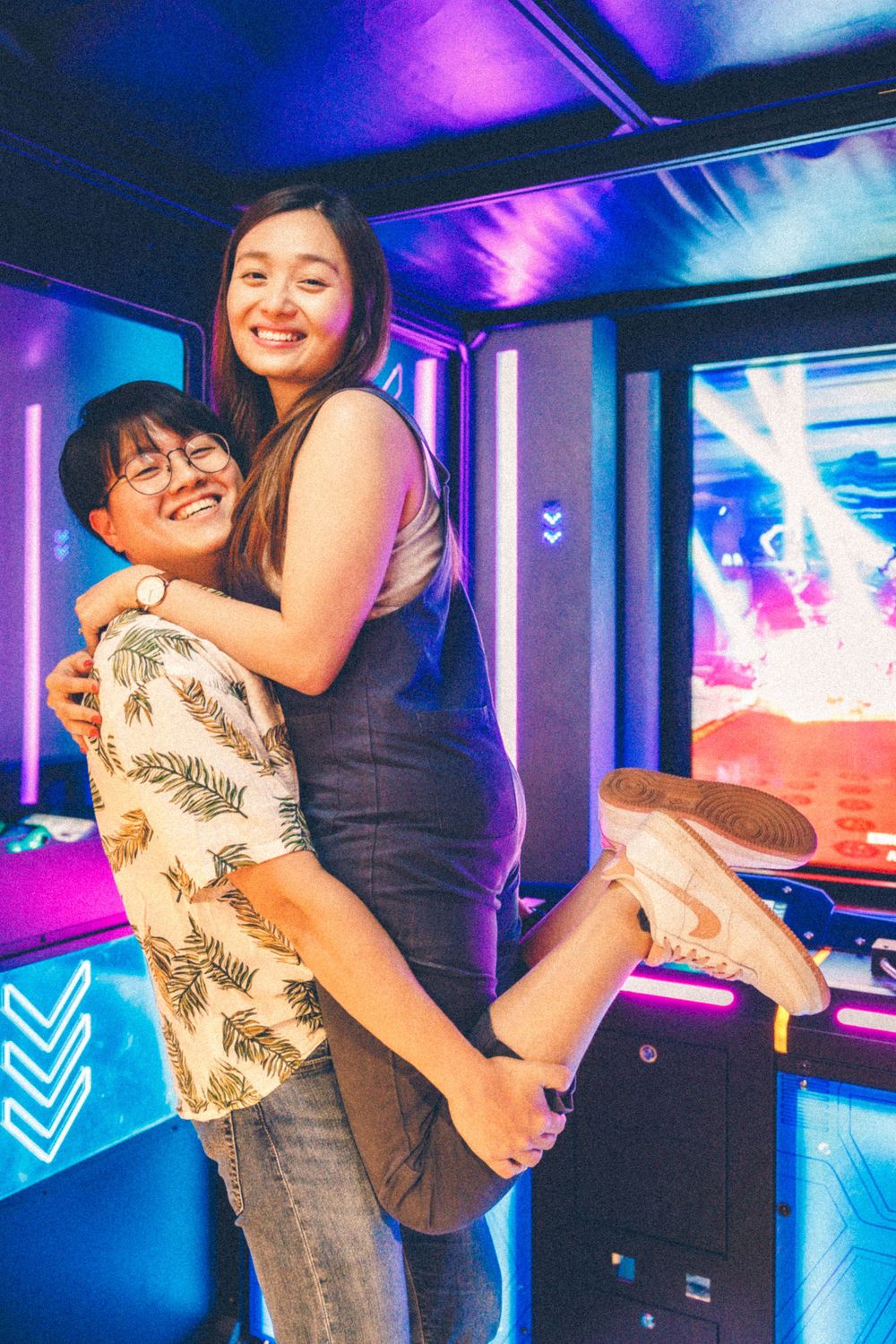 A joyful couple hugs and poses playfully in a neon-lit arcade environment.