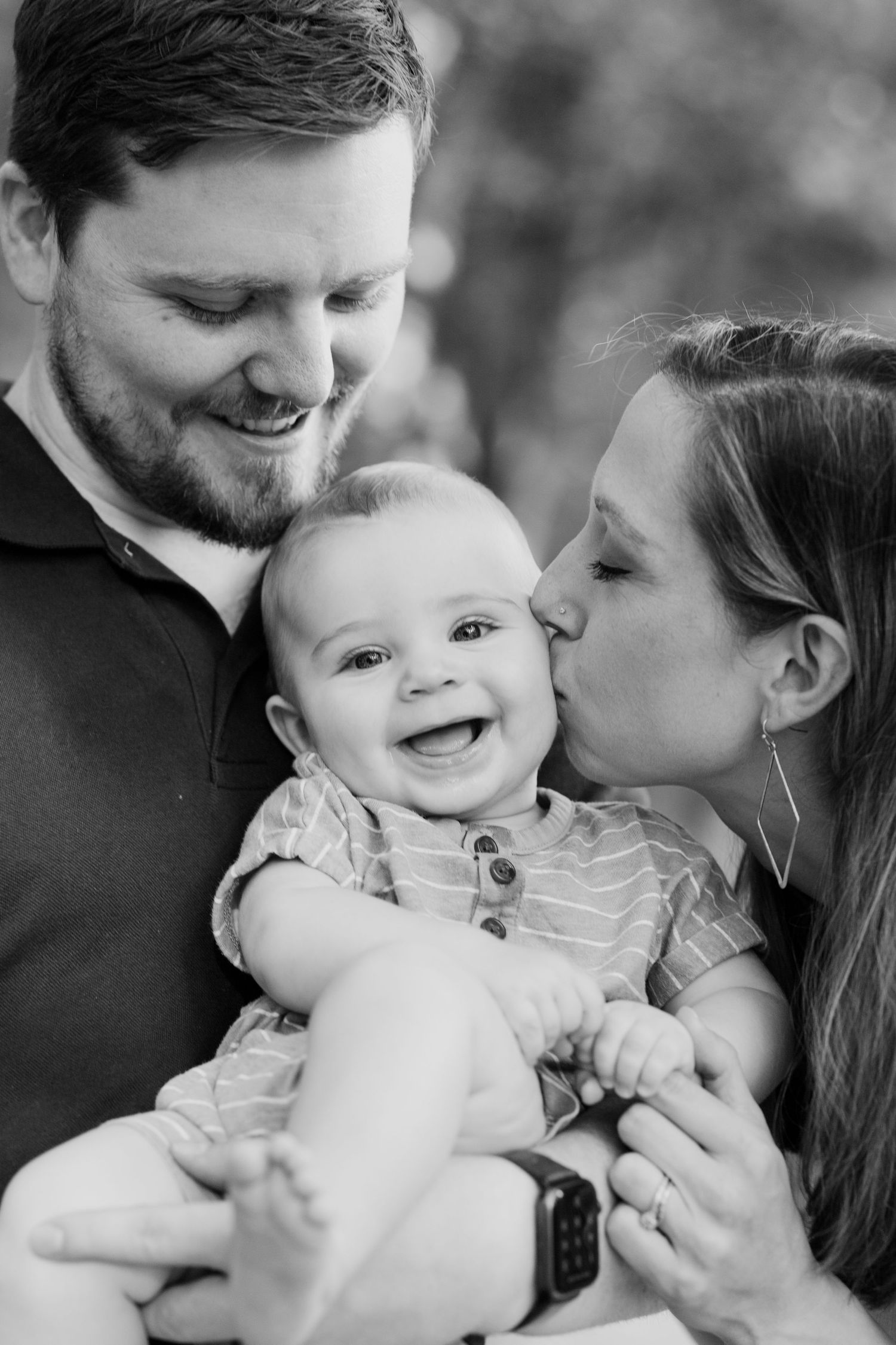 Black and white portrait of parents sharing a tender moment with smiling baby.
