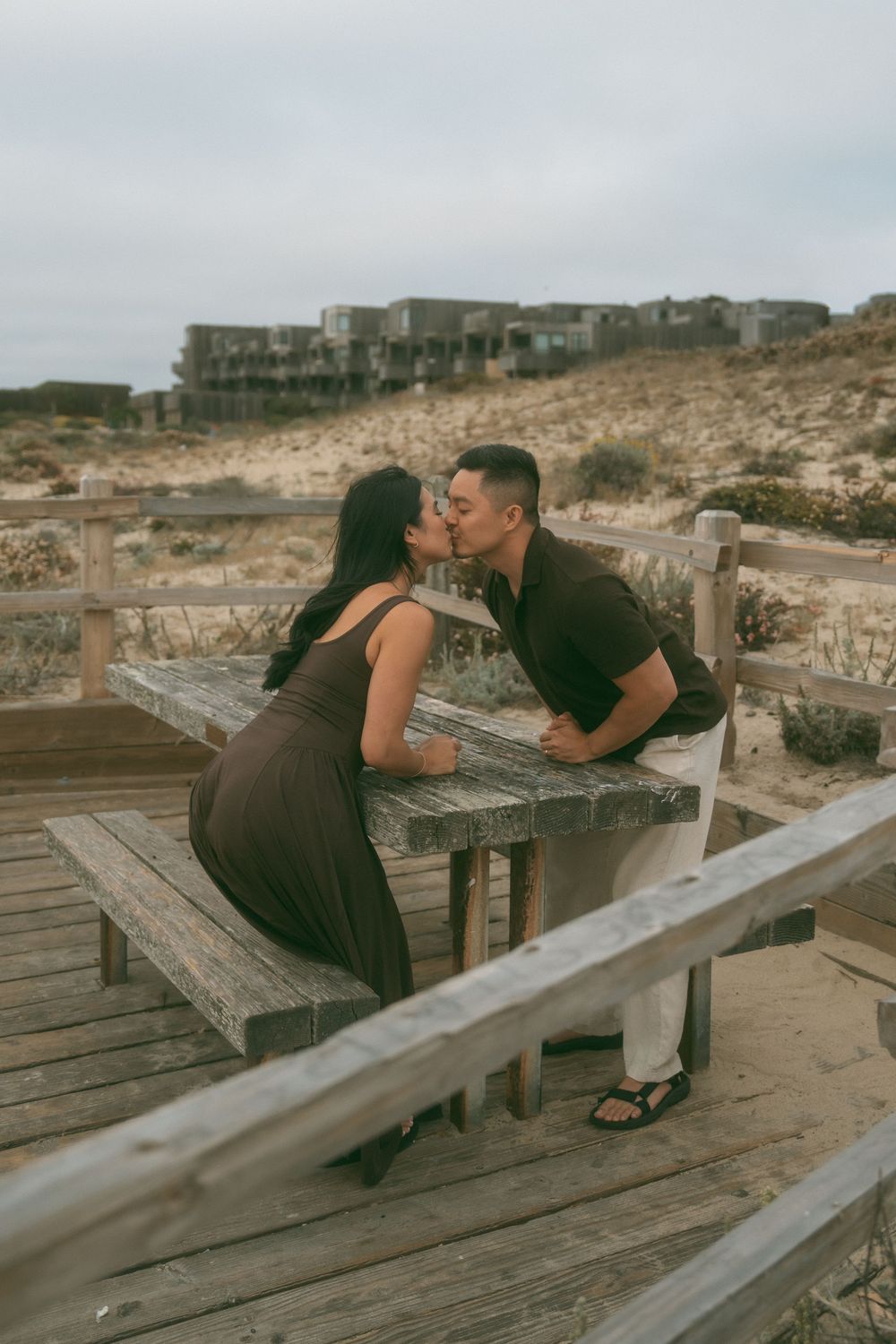 Couple sharing a romantic kiss at a weathered wooden beach picnic table with sandy dunes and buildings in the background.