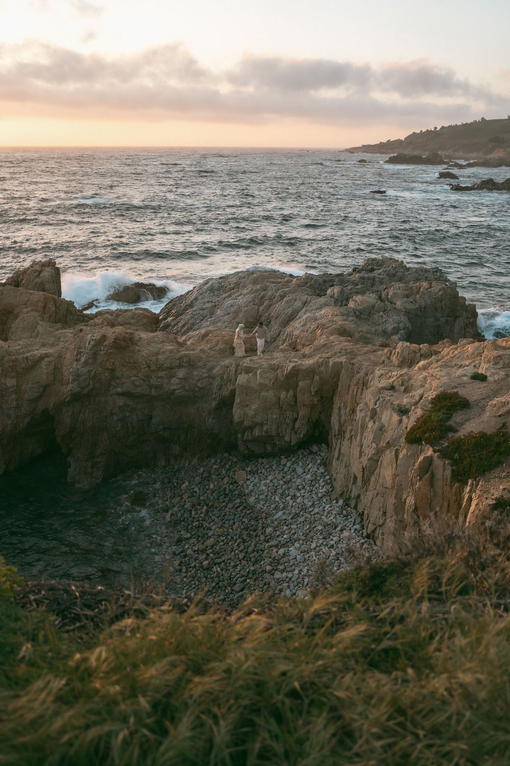Couple standing on rocky coastal cliffs at sunset with ocean waves and warm golden sky in the background.