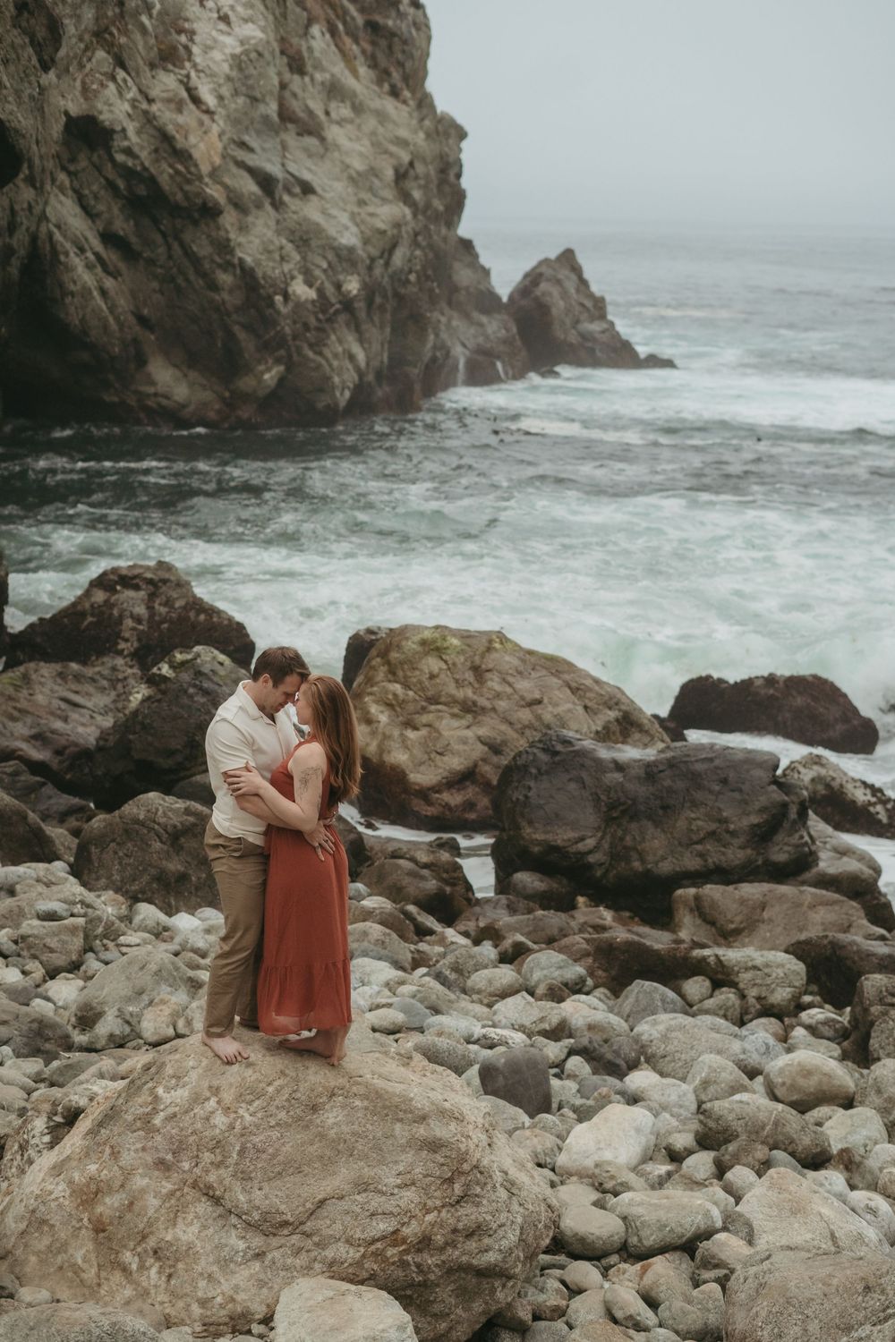 A couple embracing on rocky coastal cliffs with crashing waves and misty ocean in the background.