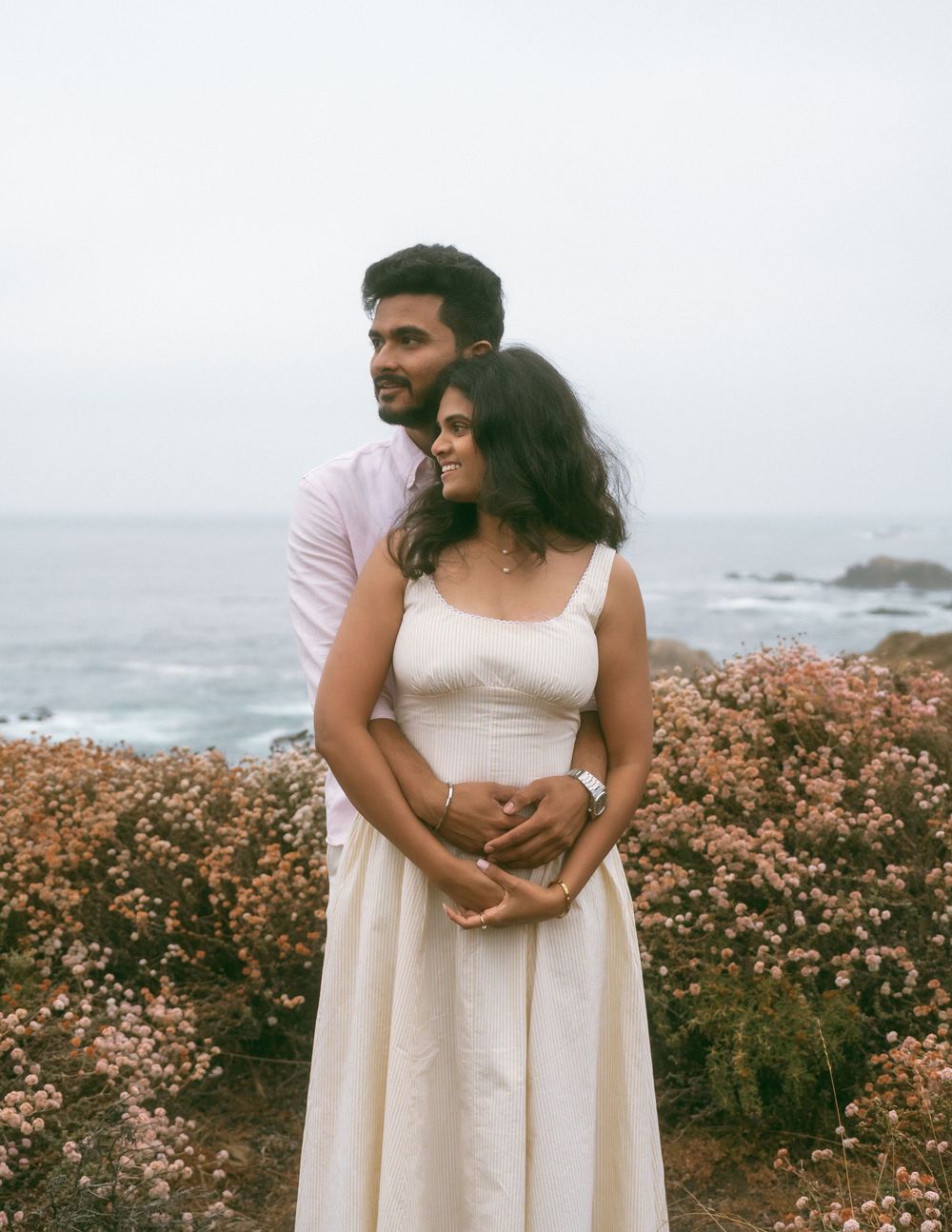 Couple embracing near coastal cliffs with wildflowers, woman in white dress, man in pink shirt, ocean backdrop.