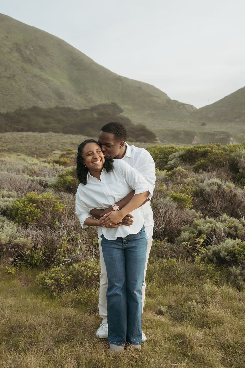 A couple embraces in a green hillside meadow, the man kissing the woman's cheek as she smiles joyfully.