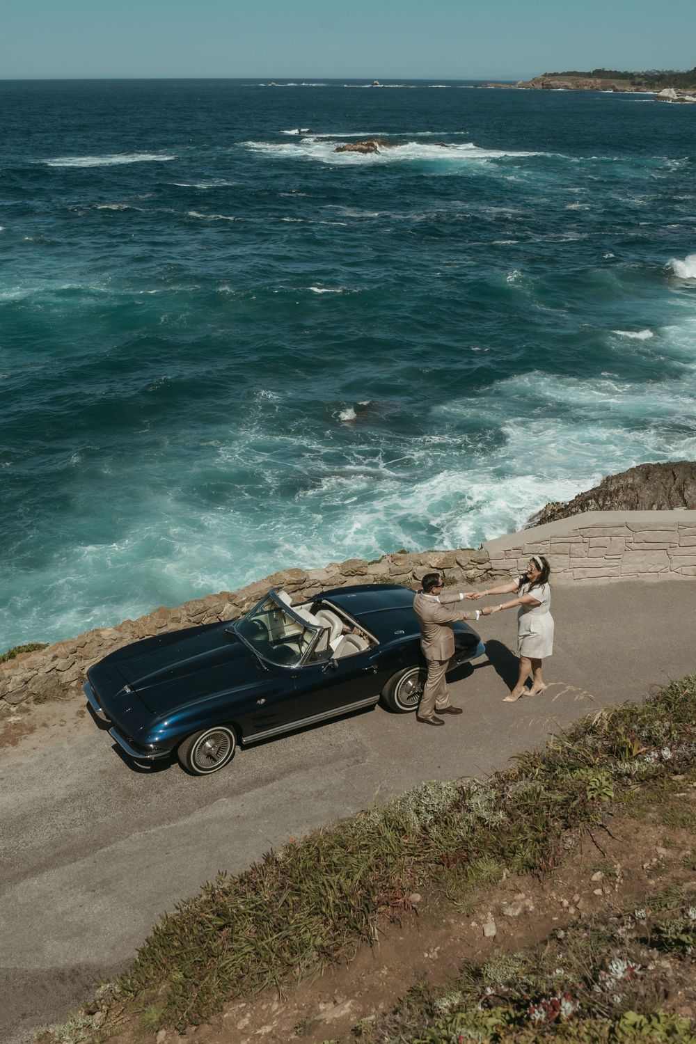 Couple holding hands beside a vintage blue convertible on a coastal cliff overlooking turquoise ocean waves.