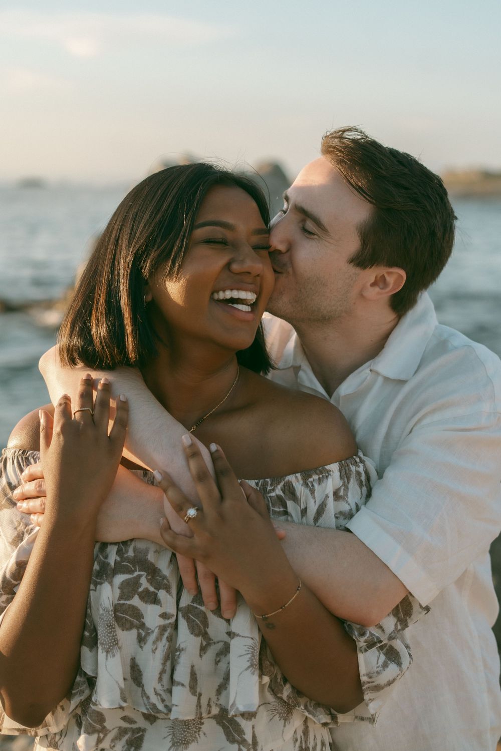 A joyful interracial couple embracing on a beach at sunset, woman laughing as man kisses her cheek.