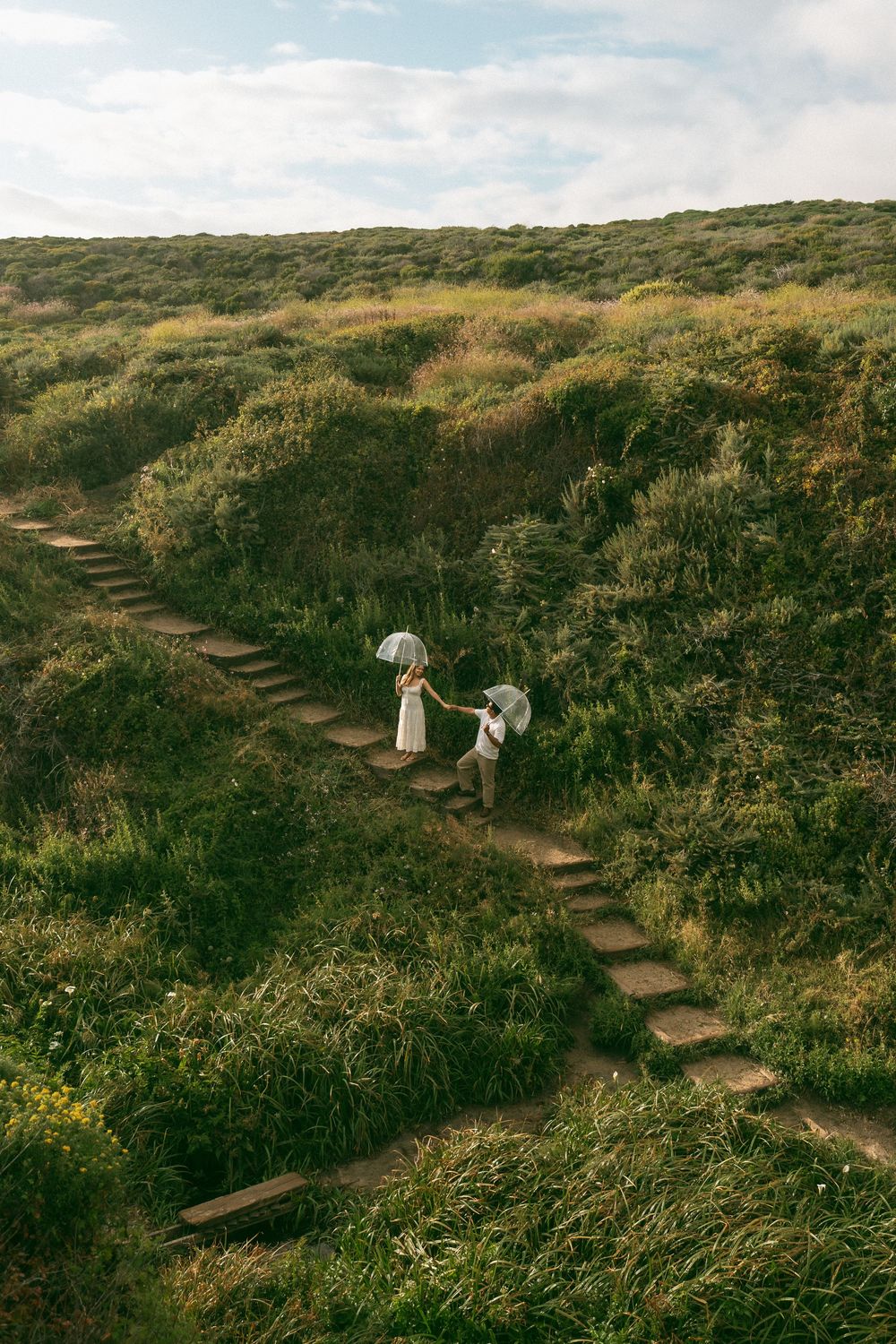 Couple holding umbrellas climbing stone steps through lush green hillside, romantic outdoor wedding photo.