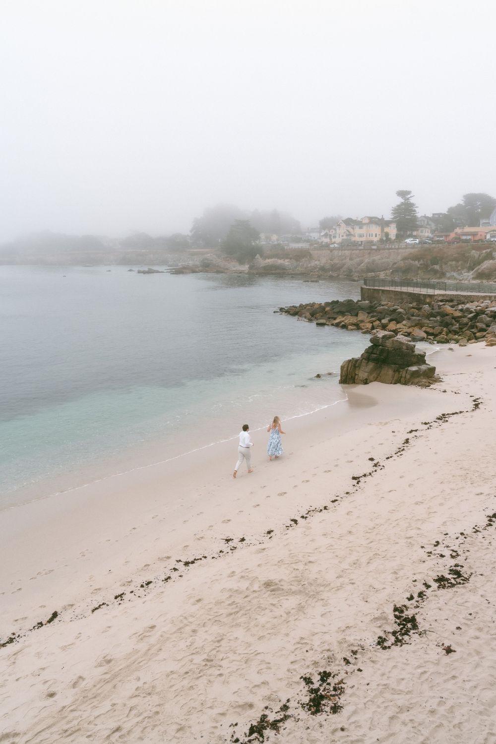 Couple walking hand in hand on a misty white sand beach with calm turquoise water and coastal town in background.