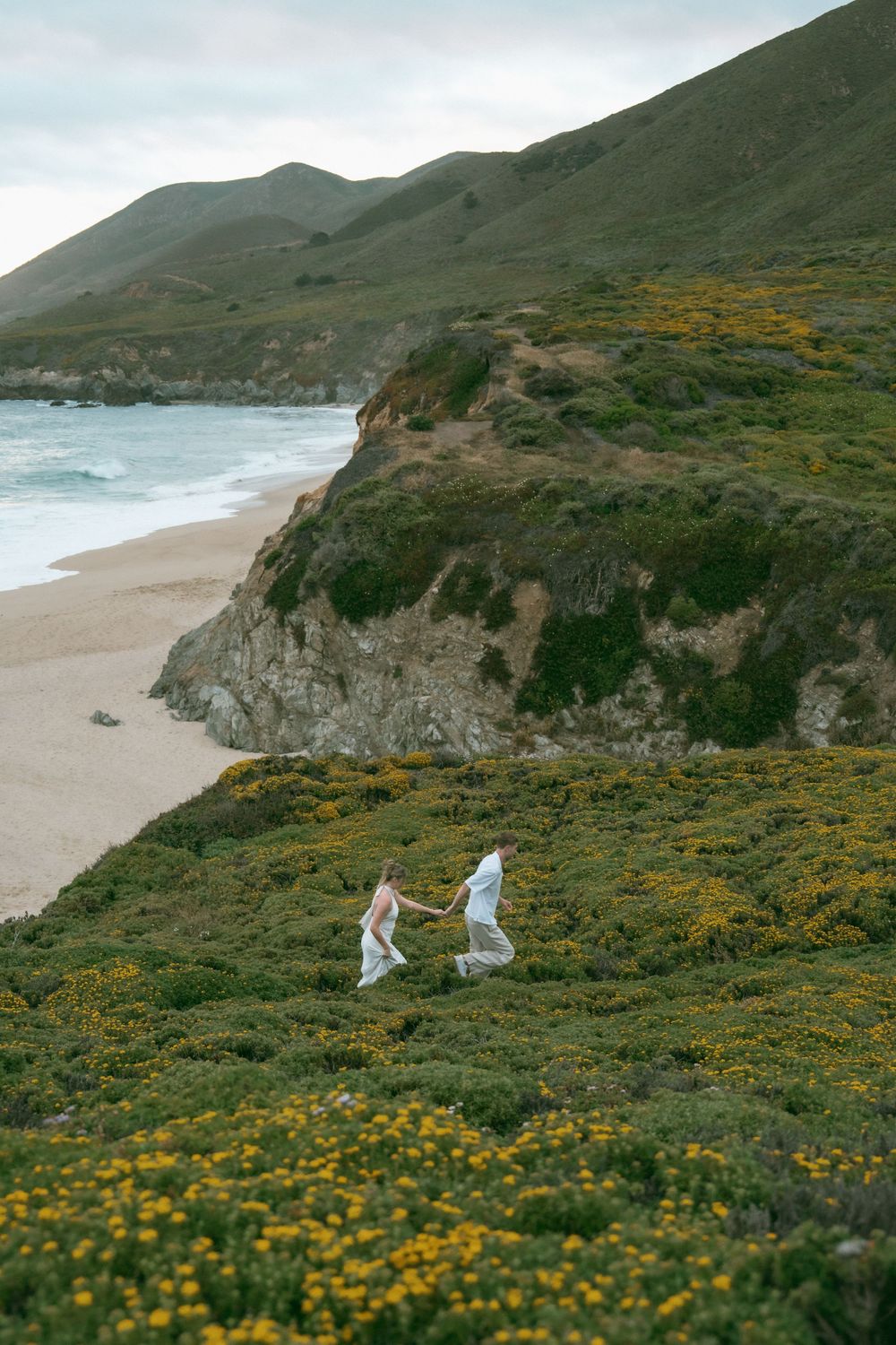 Couple in white holding hands running through yellow wildflowers on a coastal cliff overlooking a sandy beach in Big Sur.