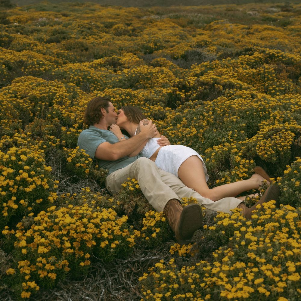 Romantic couple kissing while lying in a vibrant field of yellow wildflowers outdoors.