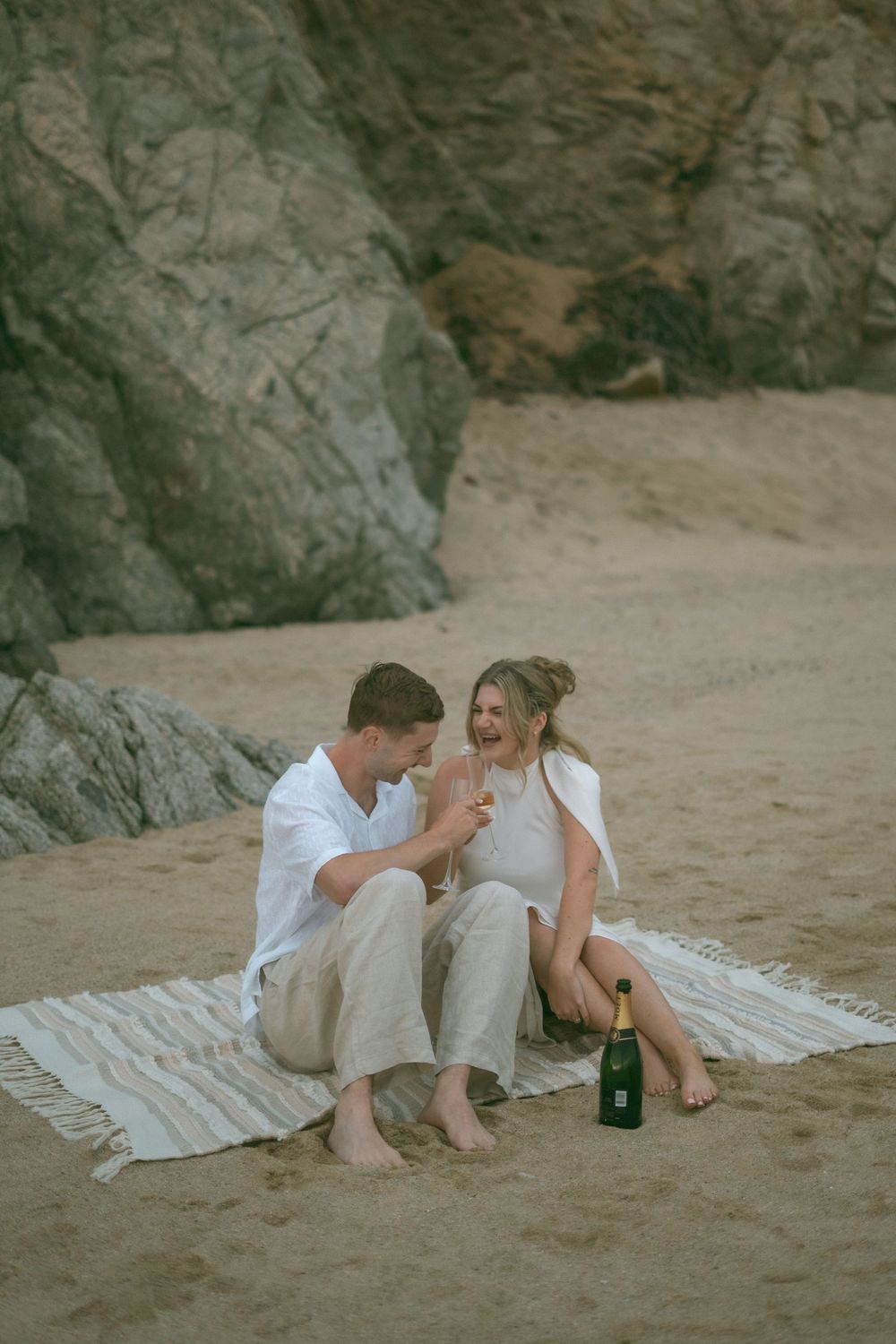 Couple sitting on a blanket on the beach, toasting with champagne glasses near large rocks.