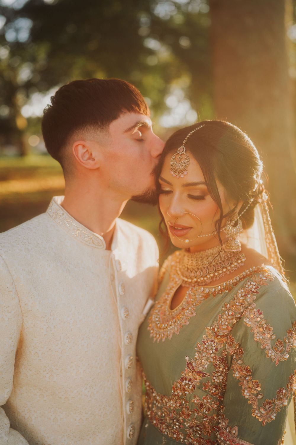 An intimate wedding portrait showing a tender forehead kiss in golden sunset lighting.