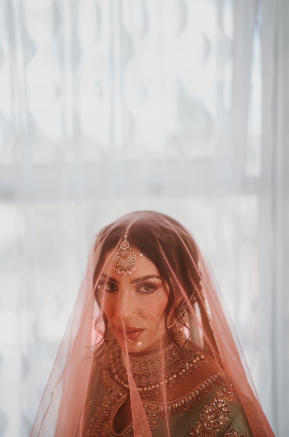 An Indian bride in traditional red veil and intricate gold jewelry shown in soft romantic lighting.