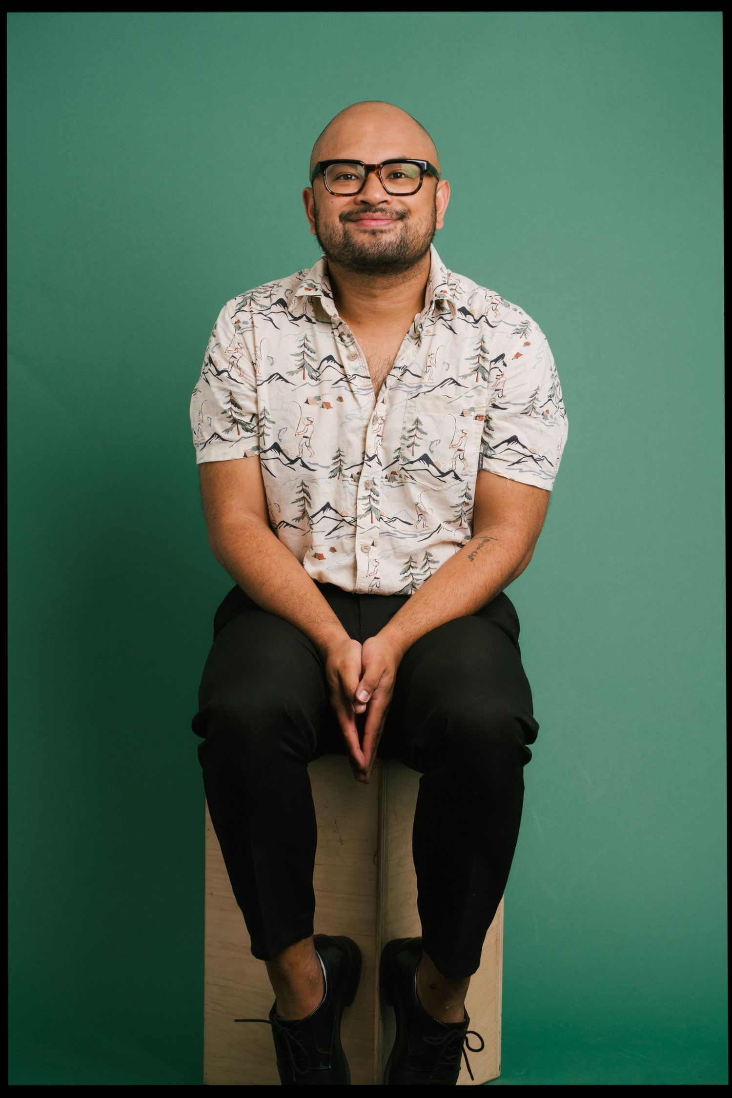 Portrait against a green backdrop of someone wearing glasses, a patterned short-sleeve shirt and black pants.