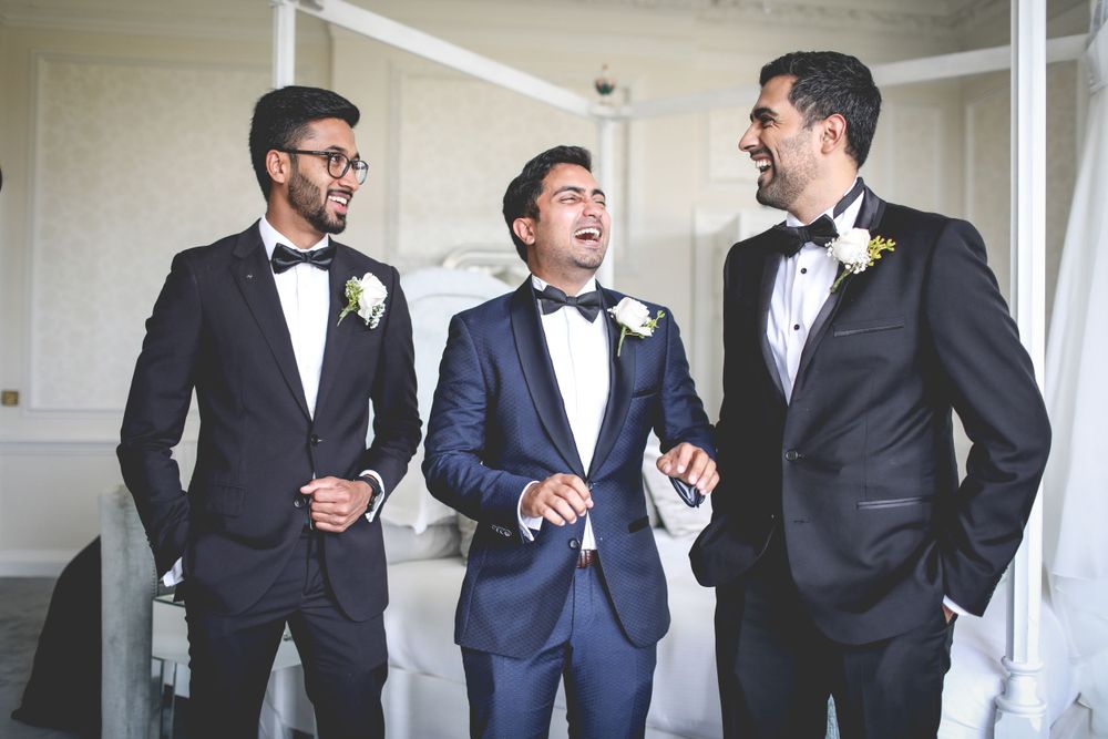 Three groomsmen in tuxedos sharing a candid moment and laughing before the ceremony. Hindu Wedding Photography