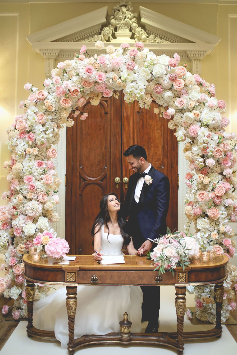 Wedding ceremony setup with luxurious floral arch of pink and white roses framing an elegant wooden table. Hindu Wedding Photography
