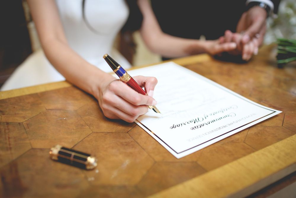 Close-up of hands signing a marriage certificate with a fountain pen on wooden table. Hindu Wedding Photography
