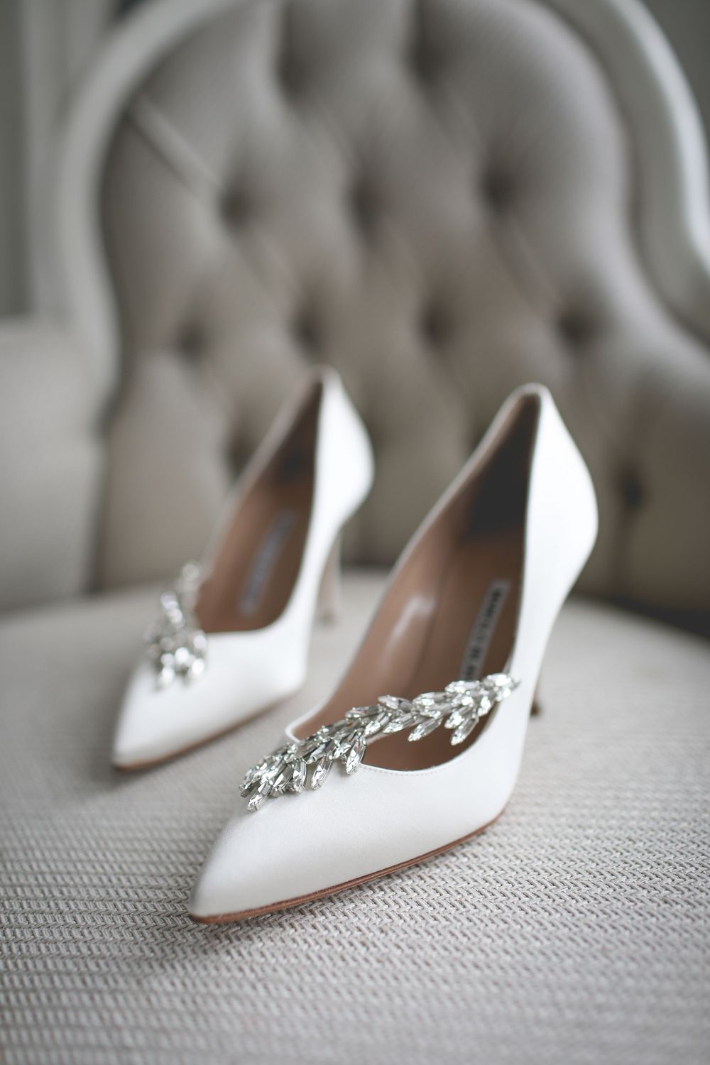 Elegant white bridal shoes with crystal embellishments displayed against grey tufted chair. Hindu Wedding Photography